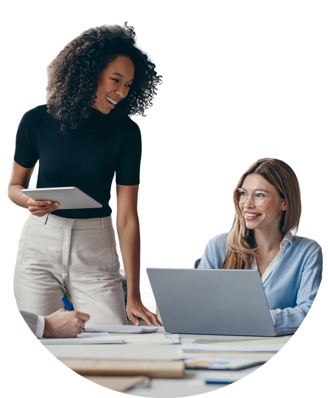 Two smiling woman in business casual discuss business. The woman on the left is holding a tablet and the woman on the right types on a laptop.