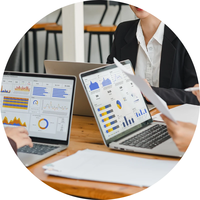 People in business attire sit around a table with laptops displaying colorful charts and graphs, reviewing printed reports together during a meeting.