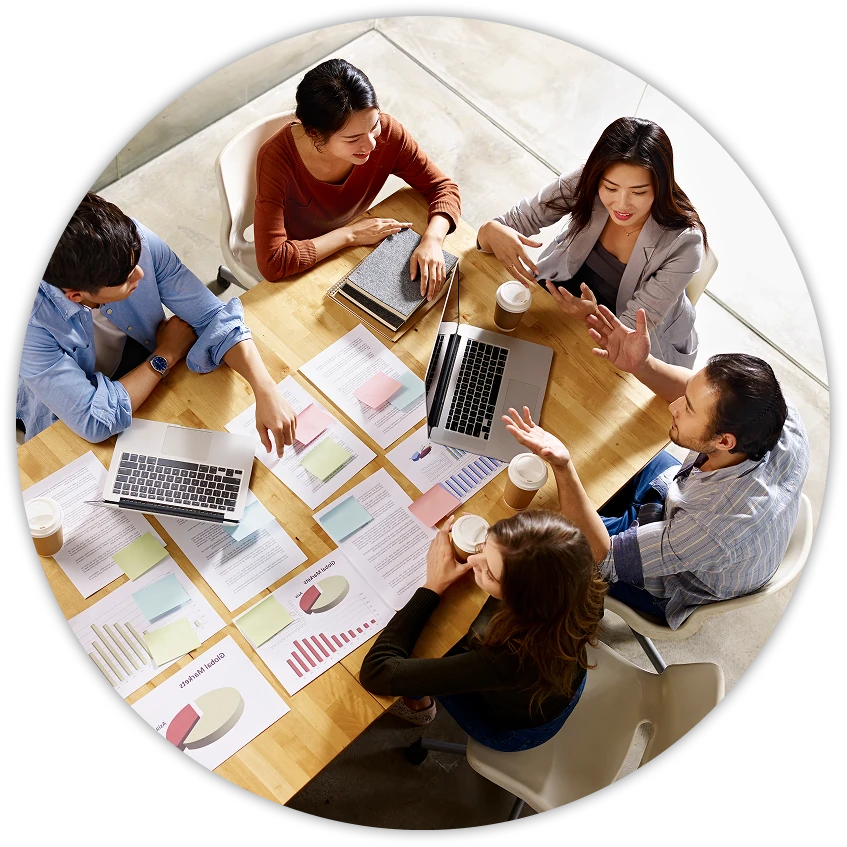 Overhead view of five coworkers in a meeting around a wooden table with two laptops, printed charts and reports covered in sticky notes, and several coffee cups as they talk and gesture.