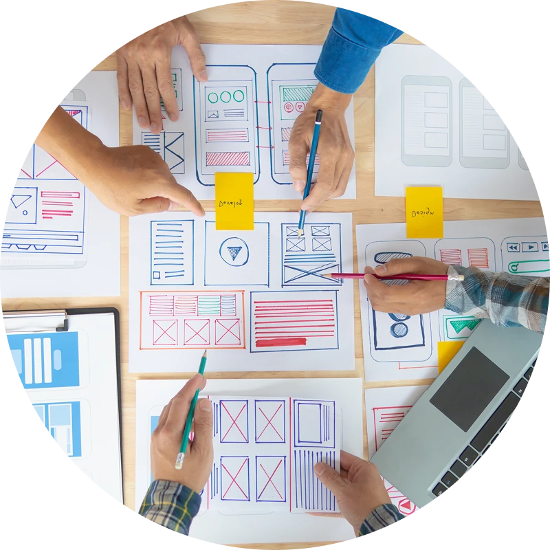 Four people’s hands work together over a table covered with hand-drawn mobile app wireframes and layout sketches. They point and draw on the papers with colored pens, with sticky notes and an open laptop nearby, suggesting a collaborative UX design or website planning session.
