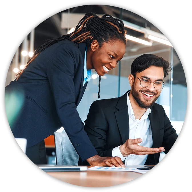 Two colleagues in business attire smiling and collaborating at a desk, with one standing and pointing at something on the table while the other sits and gestures as they talk.